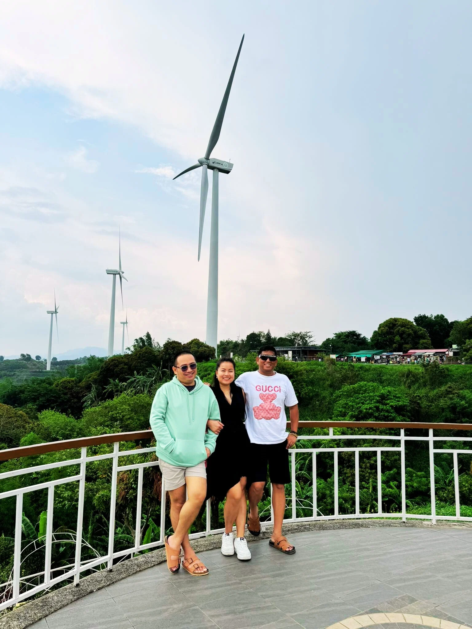 Three visitors posing at the viewing deck railing at Pililla Wind Farm in Rizal, with a tall wind turbine behind them