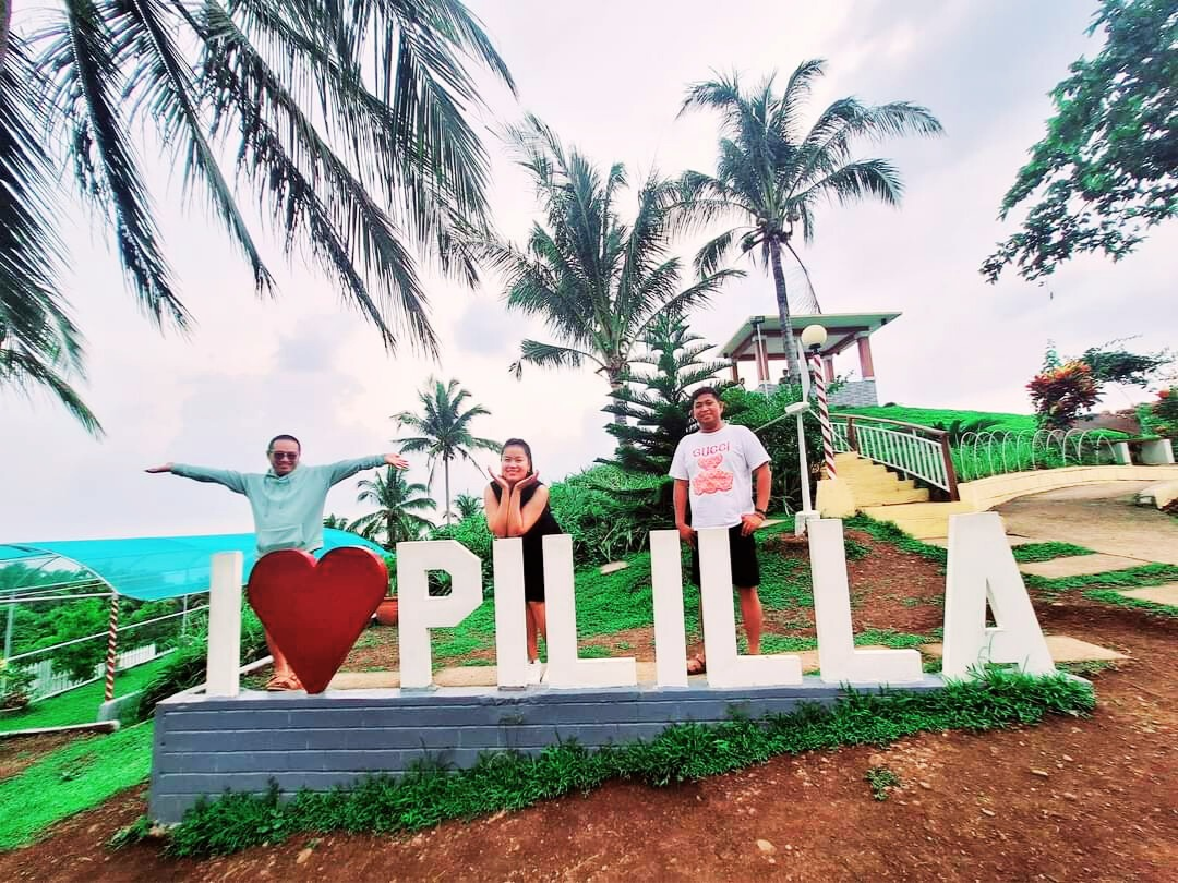 Three visitors posing at the I Love Pililla sign near Pililla Wind Farm, with palm trees and the viewing area behind them