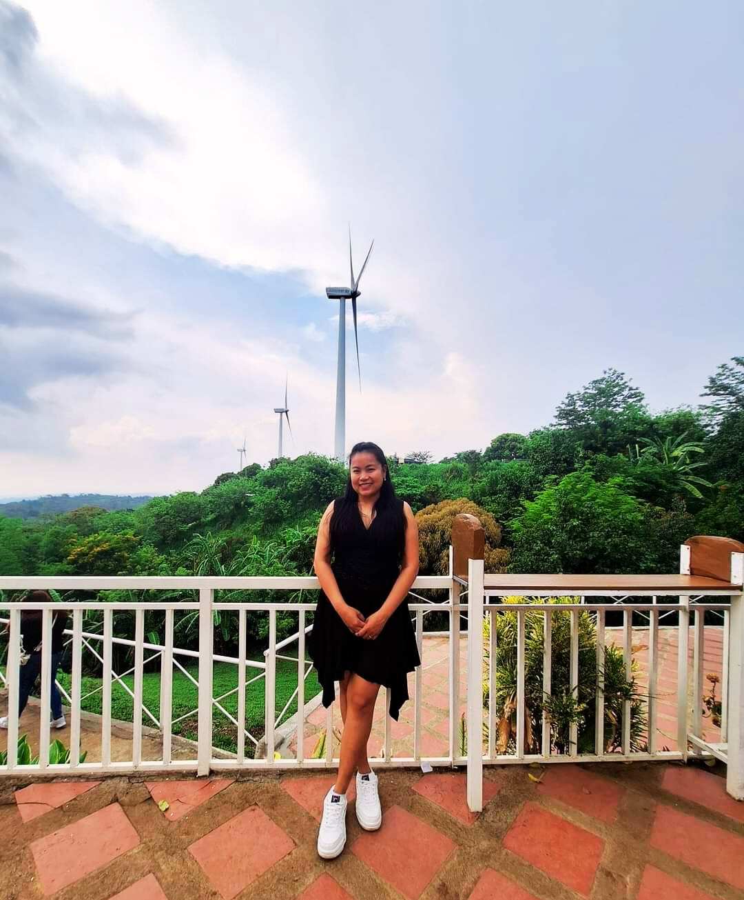 A visitor standing at the viewing deck railing at Pililla Wind Farm in Rizal, with a tall wind turbine and green hills behind her